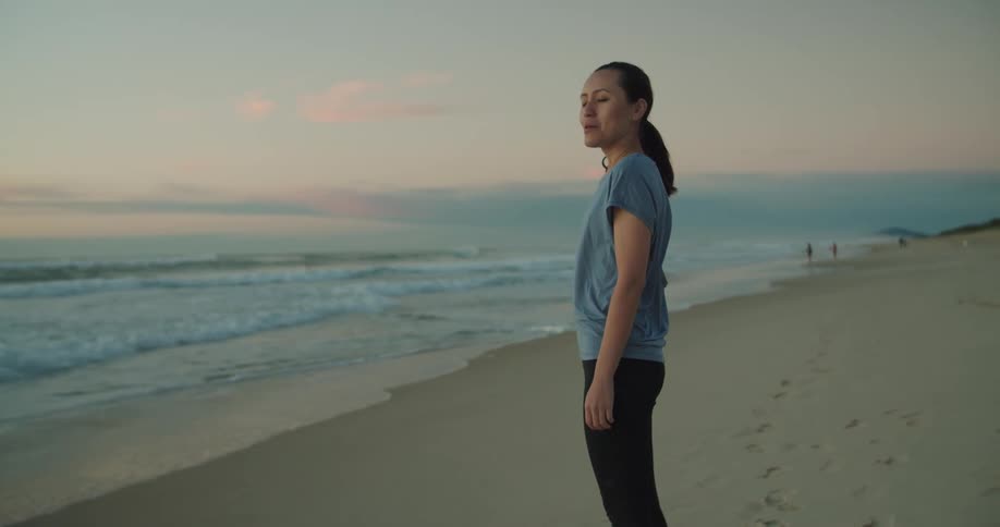 A woman stands on the beach and takes a deep breath and smiles as she watches the waves roll in