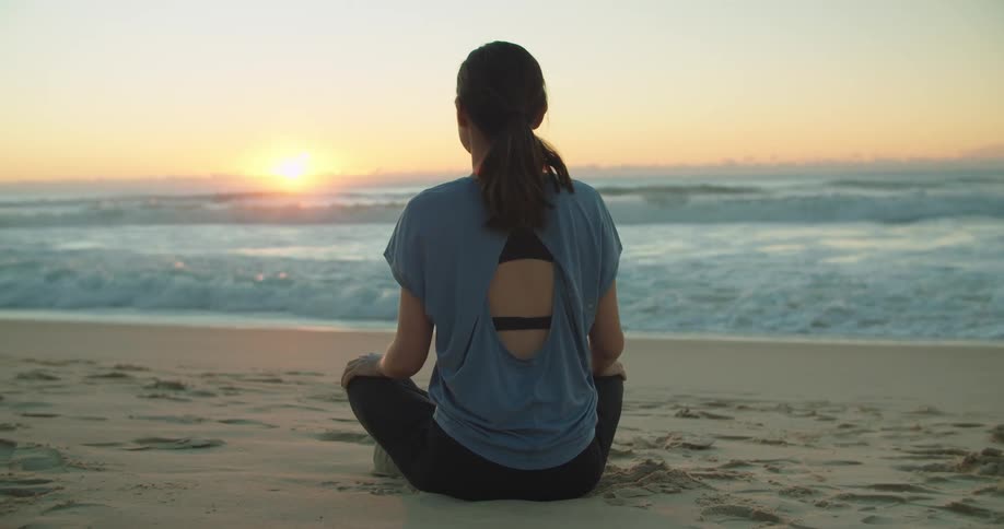 Woman sitting on the sand of a beach next to the ocean at sunset