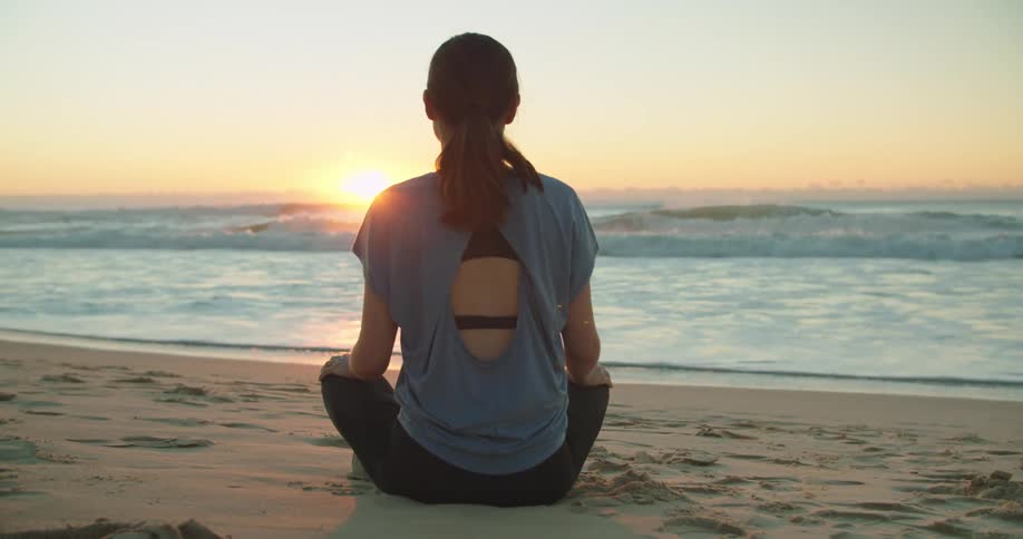 A woman sits on the sand looking out at the ocean waves with the sun on the horizon