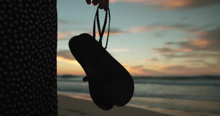 Silhuette of a woman holding a pair of sandals on the beach at sunset