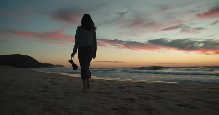 A woman walking barefoot alone on a sandy beach at sunset