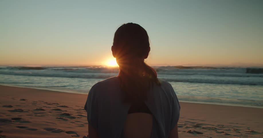 A female stands on the beach and looks out at the ocean as the sun is barely peeking over the horizon