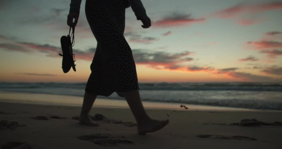 Woman walking barefoot across the beach is silhouetted against the red sky