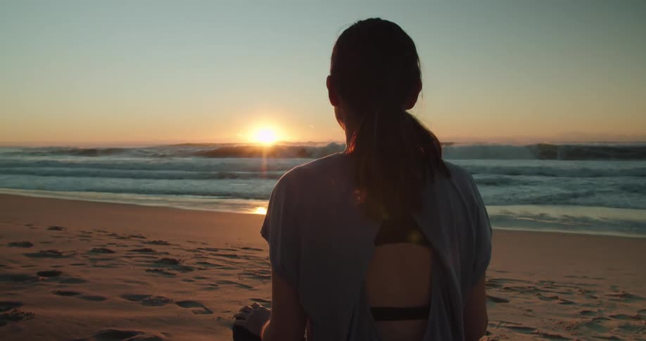 A woman sits cross legged and faces the ocean while meditating on the beach