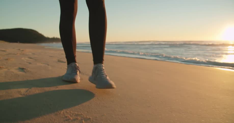 Legs in leggings and sneakers walk across beach sand as the sun glows on the horizon over the ocean