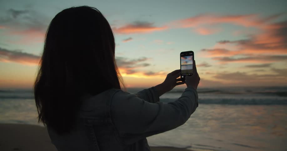 Woman with brown hair takes a photograph of sea with red sky on the beach at dusk