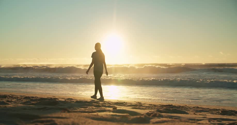 A silhouette of a woman walking on a beach with the sky and clouds in the background