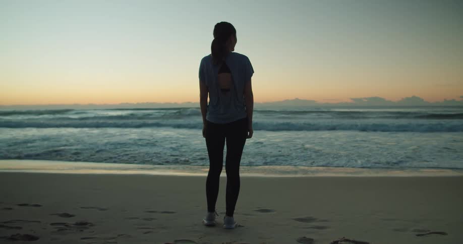 Woman stands on beach looking out to red sky on horizon over ocean