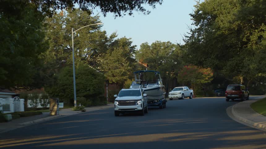 Suv pulls boat trailer carrying motorboat down street through neighborhood past houses and parked cars