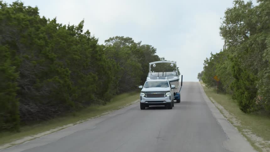 A white sport utility vehicle tows a large boat down a narrow tree lined road