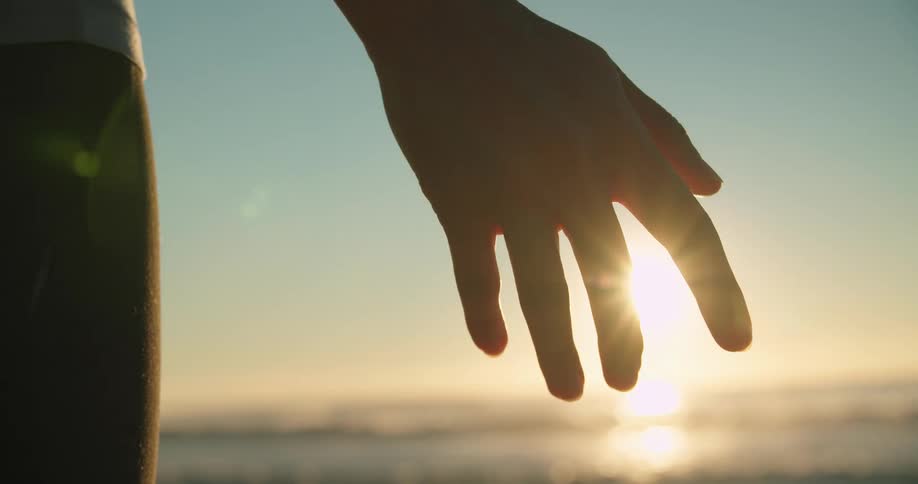 A person holds out their hand and feel the ocean breeze with their fingers as the sun is low in the sky