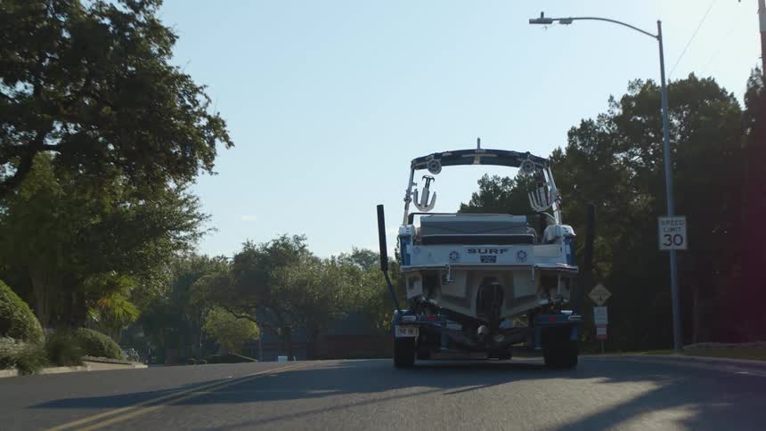 Vehicle tows trailed with motor boat along road through neighborhood passing other vehicles
