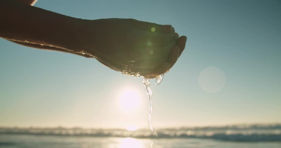 Ocean water drips from a persons cupped hands as the sun glares and shines on waves