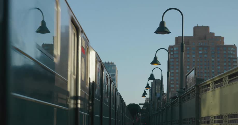 Train cars reflecting the sun move through city between building bordered by row of street lamps