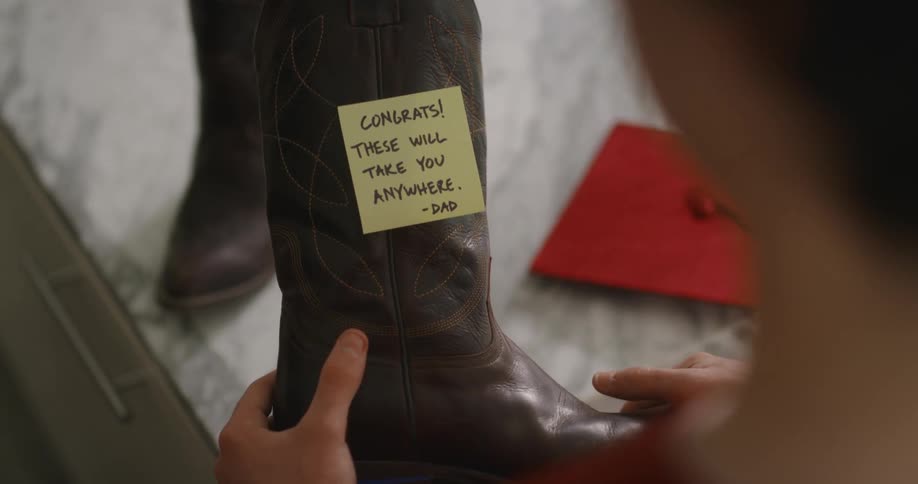 A person in a room holding a boot with a bed and a graduation cap in the background