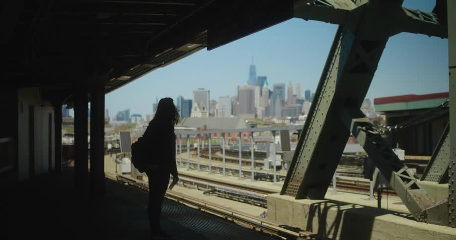 A woman with a mask and a bag standing in a train station with the buildings the sky and clouds in the background