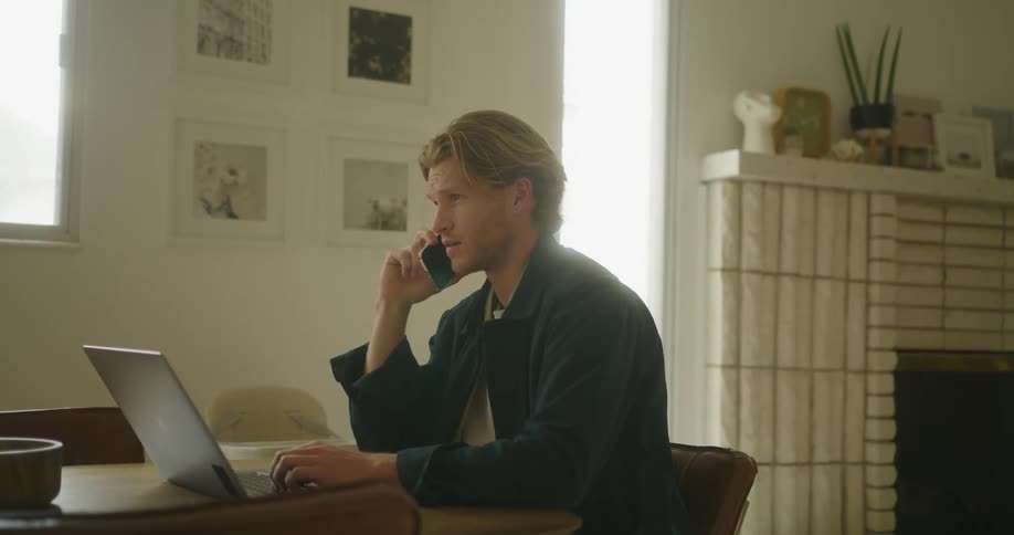 Man sitting at table with fireplace and windows nearby talks on mobile phone and uses laptop