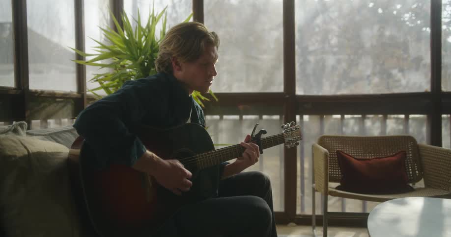 A man in a room sitting on a couch playing a guitar next to a table