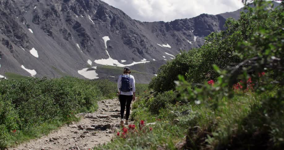 A woman wearing a backpack walks down a rocky trail next to mountains