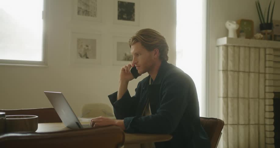 A man sits at a desk with a laptop in front of him talking on a cell phone