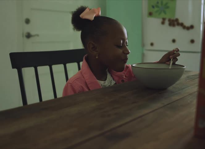 A girl in a room sitting in a chair at a table eating cereal in a bowl with a spoon
