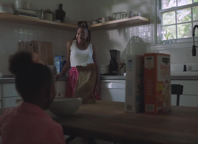 Mother stands near counter smiling at daughter sitting at wooden table eating cereal