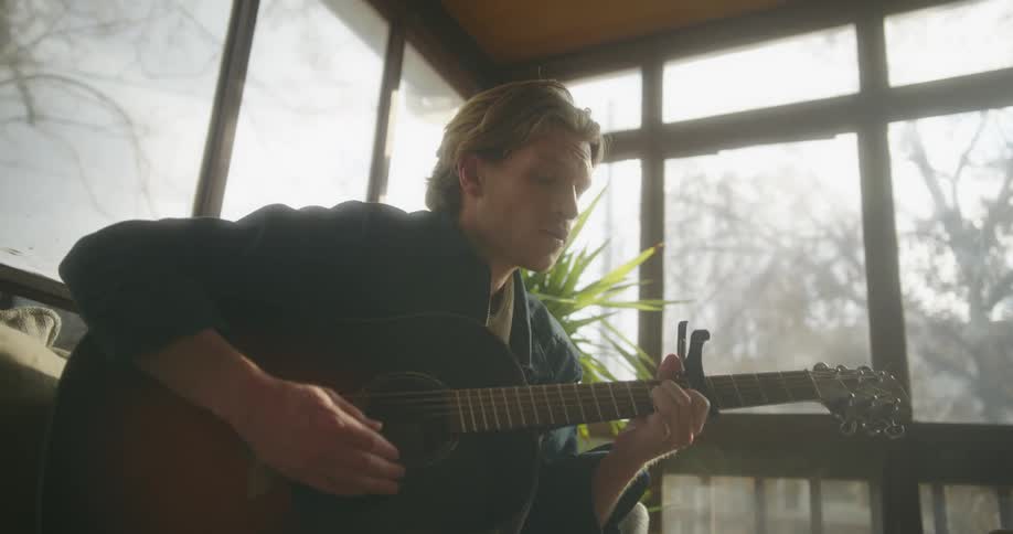 Blonde man with a black shirt plays the guitar in a room