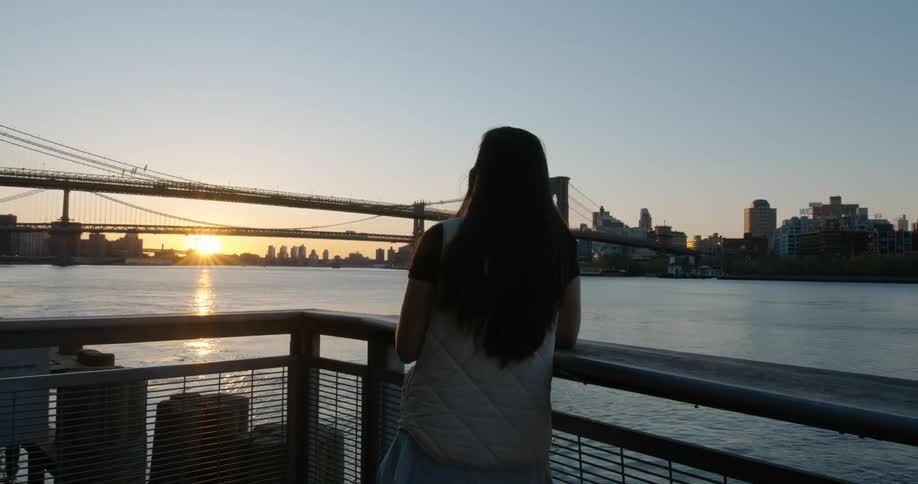 A teenage girl walks out on a patio on the shore and watches the sunset over the water