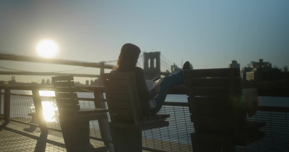 A woman sitting on a chair on a pier with a bridge buildings the sky and clouds in the background