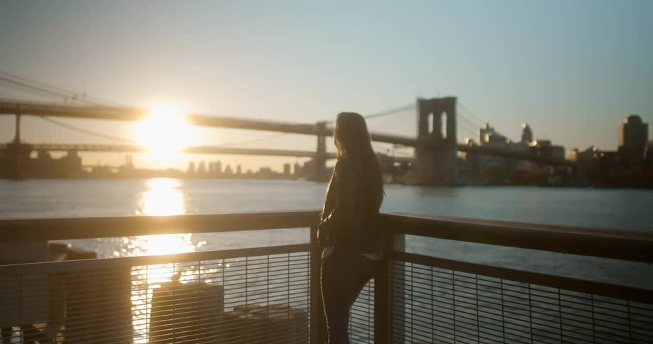 A woman with a mask and a bag walking by a pier with a bridge buildings the sky and clouds in the background