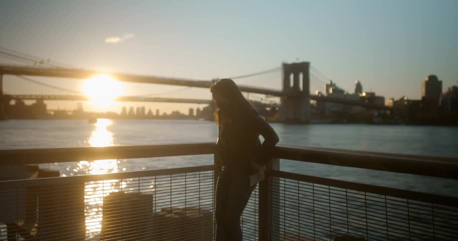 A woman with a mask standing by a pier with a bridge buildings the sky and clouds in the background