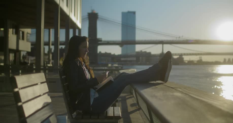 Woman sitting in a bench takes her phone out to take a picture of the river near the city
