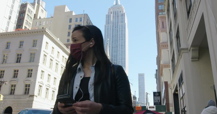 Woman wearing a covid mask holds a smart phone while wearing ear buds as she stands in the city