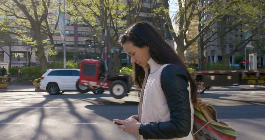 A woman with a bag walking on a city street and using a smart phone with trees and buildings in the background
