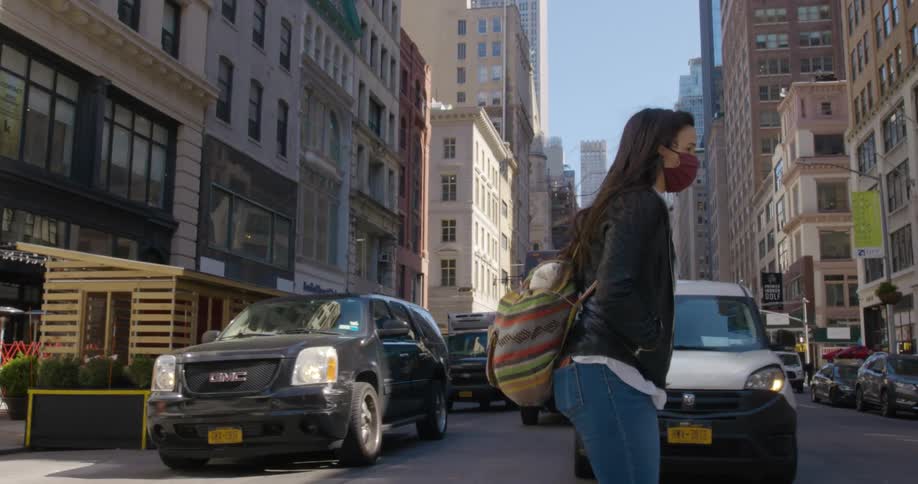 Woman with a claret red mask and black leather jacket and jeans walks across the street with vehicles and tall buildings in the background