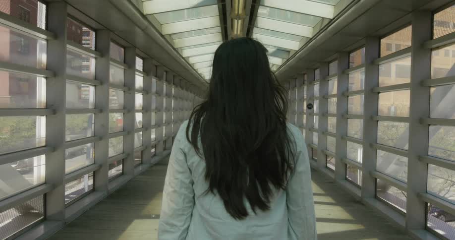 Woman walks along enclosed pedestrian walkway over street traffic with buildings visible through windows