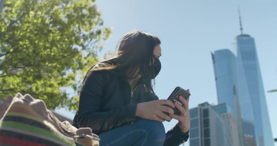 Woman with brown straight hair and a black mask a black leather jacket and jeans uses smartphone with skyscrapers in the background
