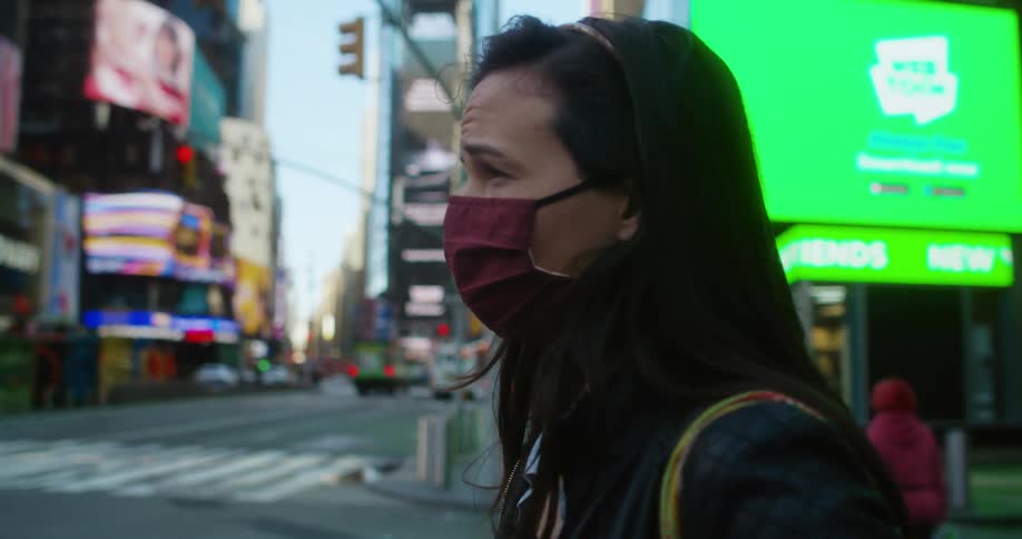 A woman wearing a cloth face covering stands on a city street with a furrowed brow looking anxious