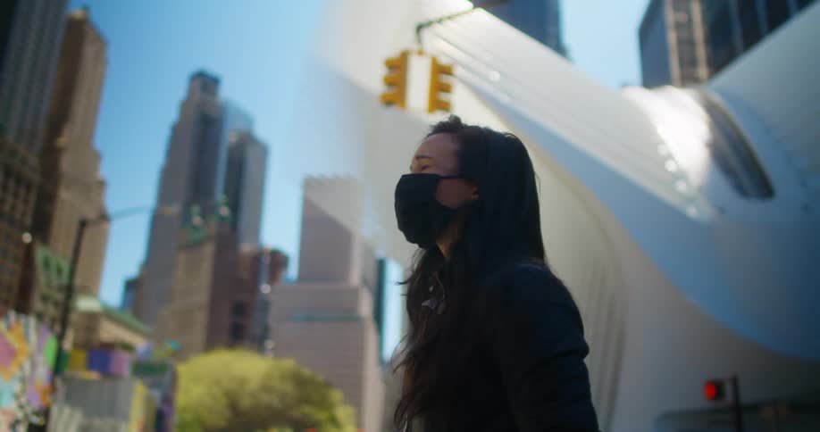 A woman with a mask walking in a city with buildings the sky and clouds in the background
