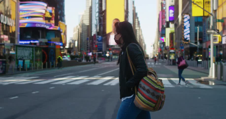 A woman with a mask crossing the street in times square with buildings the sky and clouds in the background