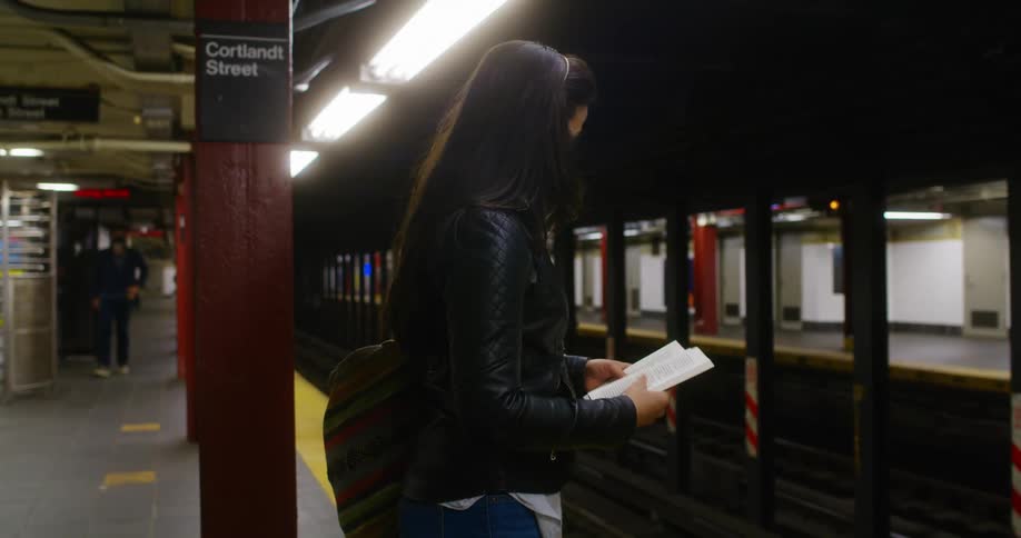 A teenage girl reads a book at a train station as a man walks on the platform
