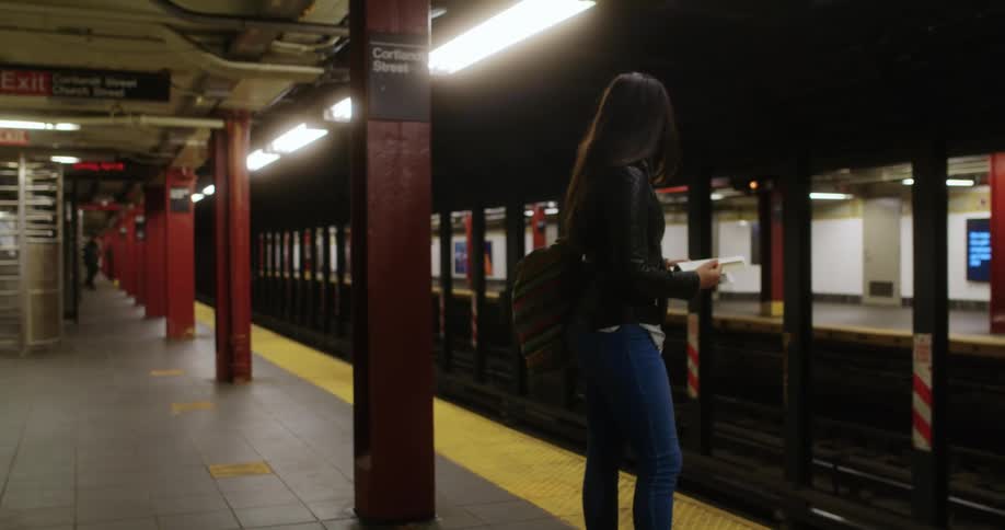 A woman with a mask and a bag standing in a subway reading a book