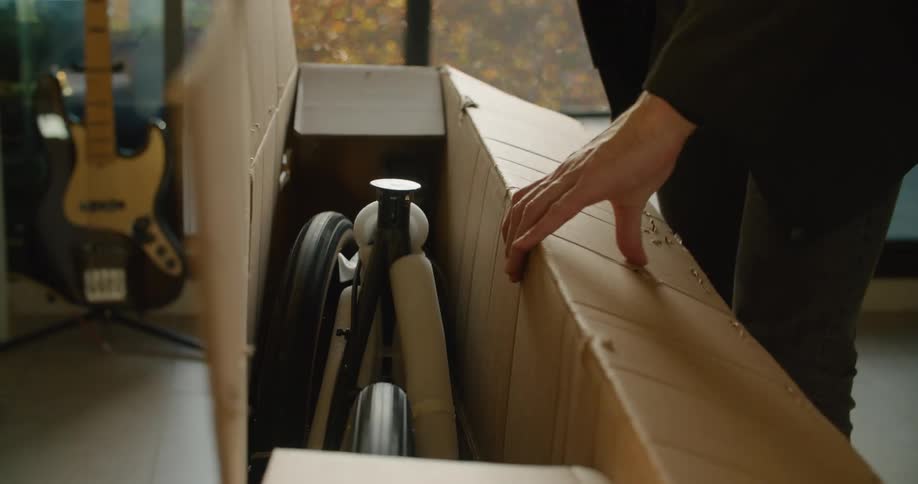 A man standing in his living room opens a tall and narrow cardboard box with electronic equipment inside