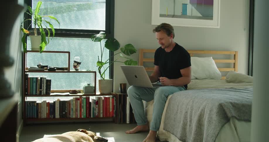 A man in a room sitting on a bed talking on a laptop with a bookshelf in the background