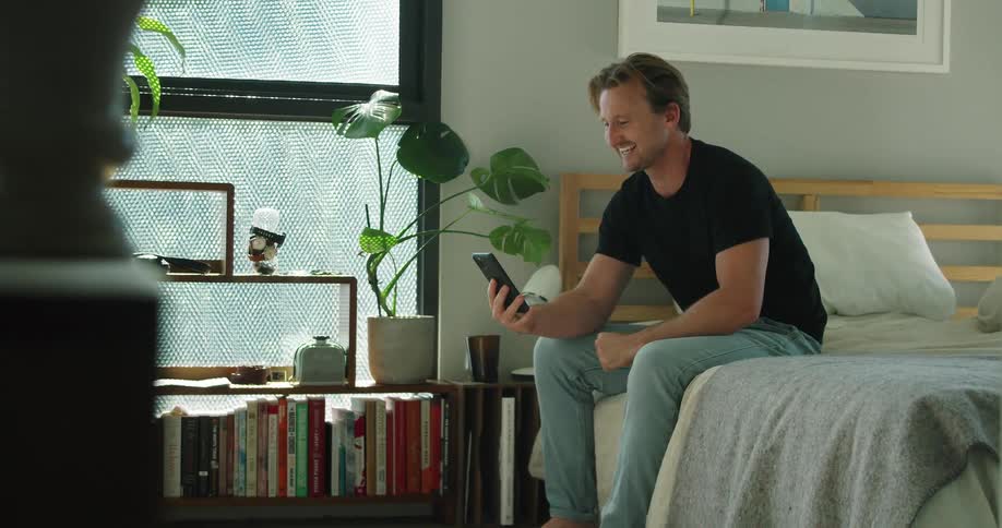 A man sitting on his bed waves at his phone as he video chats