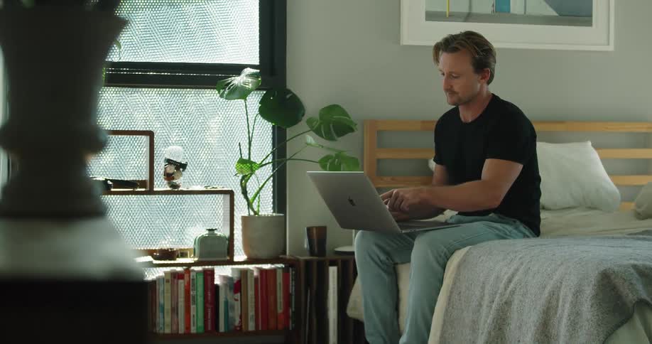 A man sits on his bed holding his laptop and participating in a video call