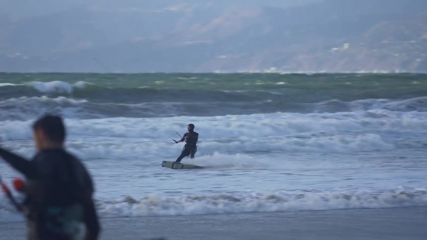 Man wearing a black wet suit kiteboarding at a beach near the mountains
