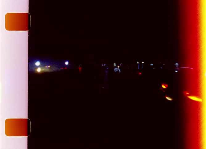 Crowd stands outside at night with cars circled around empty field