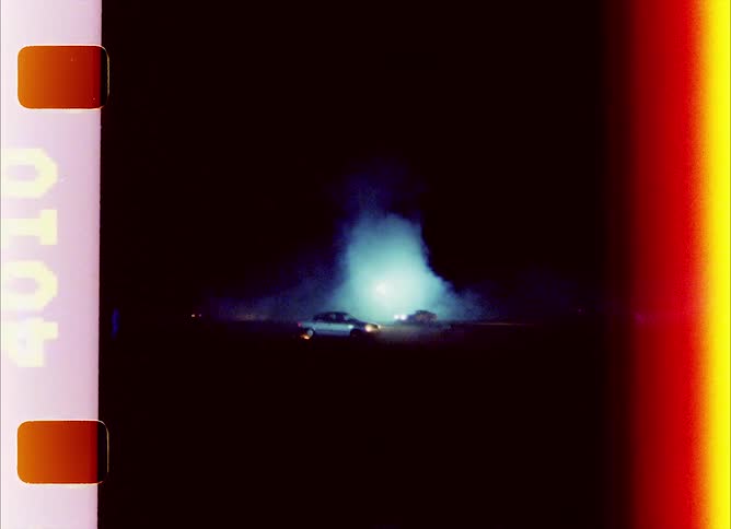 Cars driving around a track filled with white smoke at night caught on vintage film