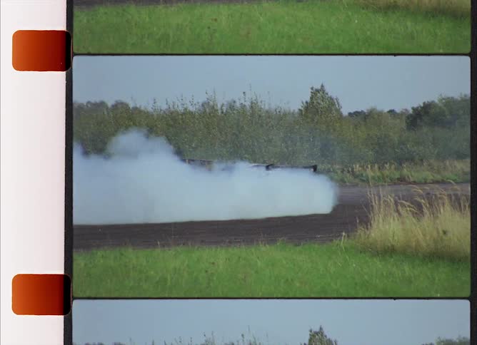 Car speeds along blacktop between fields near forest filling air with smoke under a clear blue sky